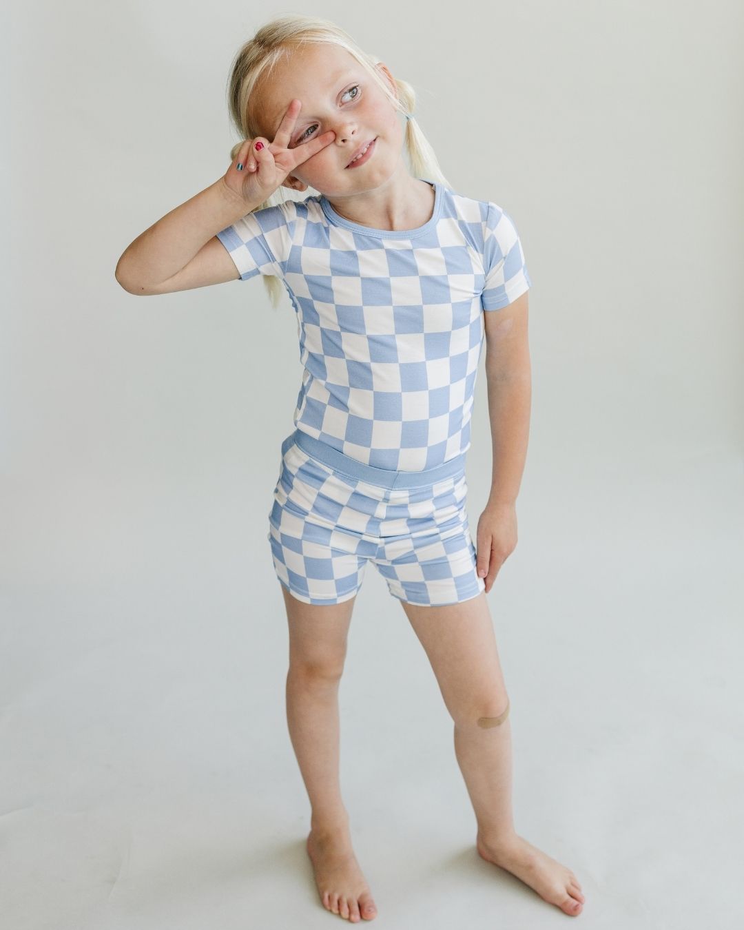 A young girl standing up and holding up a piece sign to her face. She is wearing the Lucky Panda Kids viscose bamboo two-piece short pajama set in a white and light blue checkered pattern.