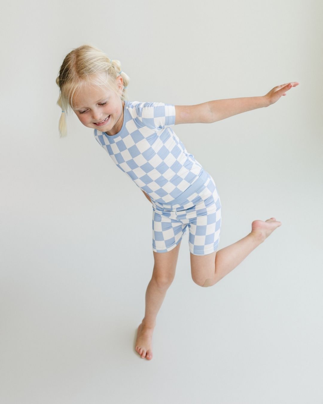 A young girl standing and lifting one leg and one arm. She is wearing the Lucky Panda Kids viscose bamboo two-piece short pajama set in a white and light blue checkered pattern.