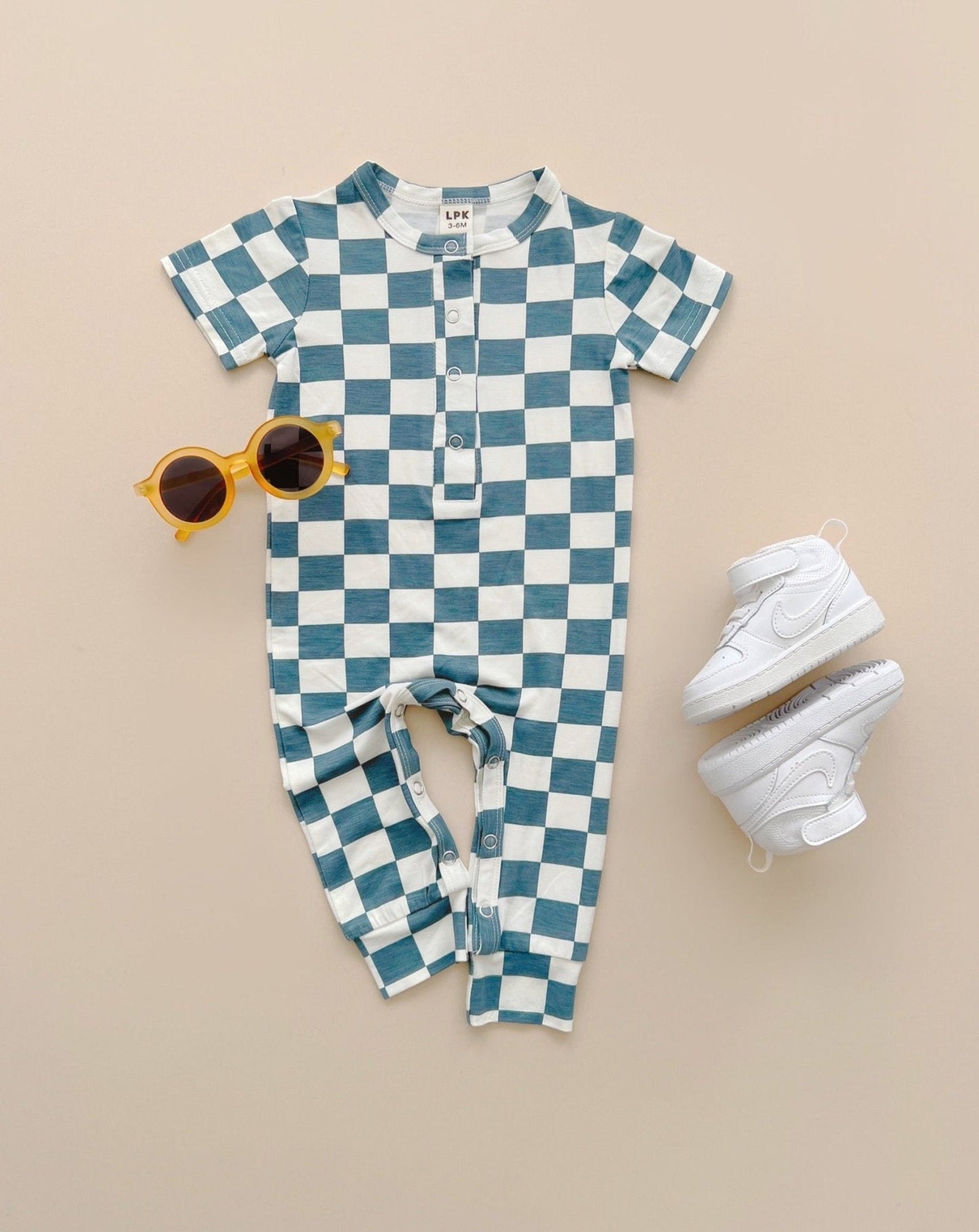 Blue and white checkered short sleeve bamboo jumsuit from Lucky Panda Kids laid out on blank background with accessories including white nike shoes and sunglasses. Sold by MilkbabyCo