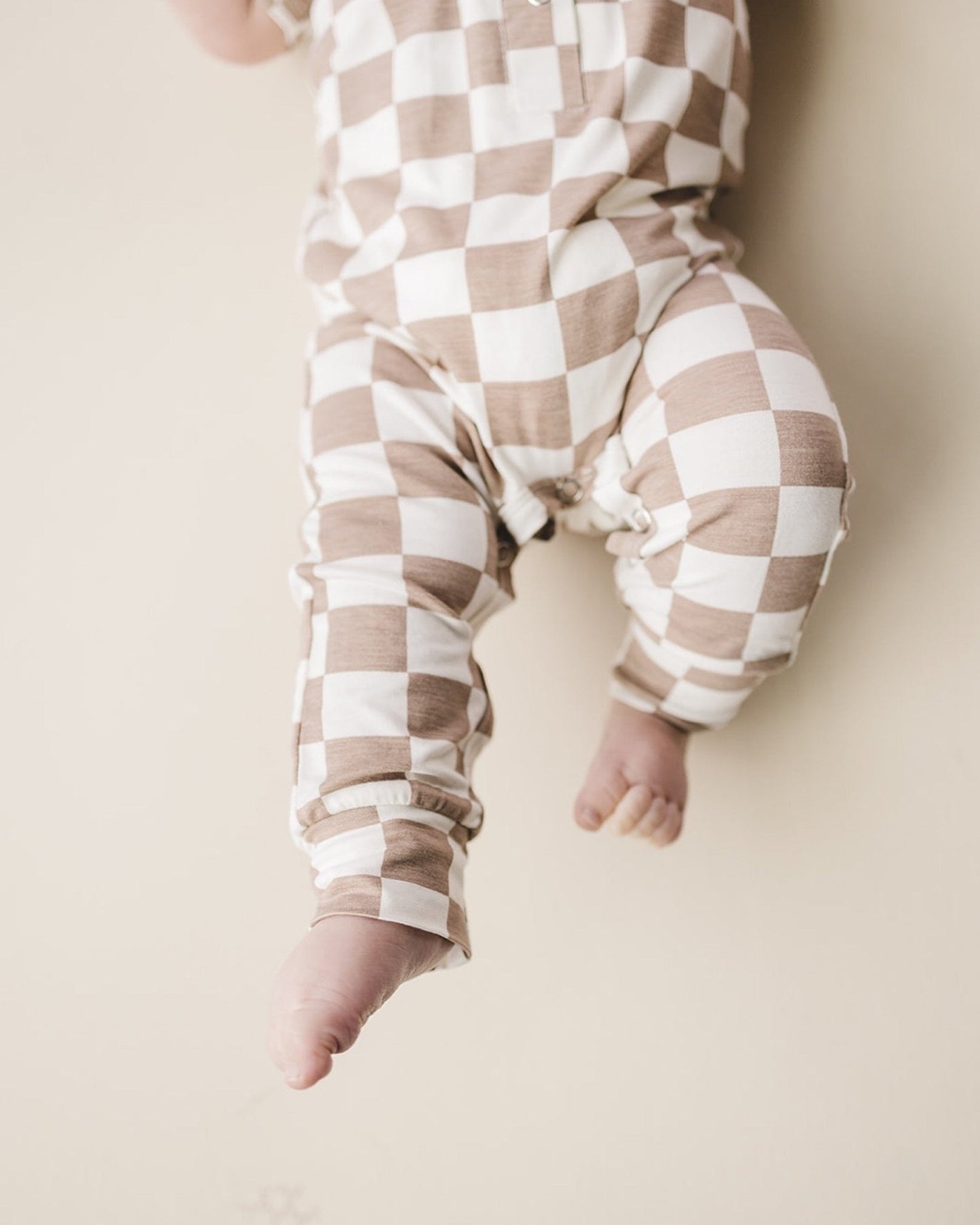 Close up lower body shot of an infant baby laying down wearing a tan and white checkered short sleeve jumpsuit from Lucky Panda Kids made from soft viscose bamboo.