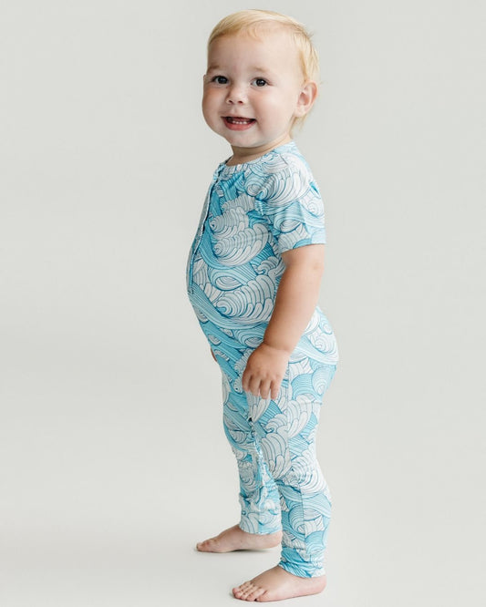 Lucky Panda Kids short sleeve romper in blue and white ocean waves design worn by a toddler boy who is standing sideways smiling. 