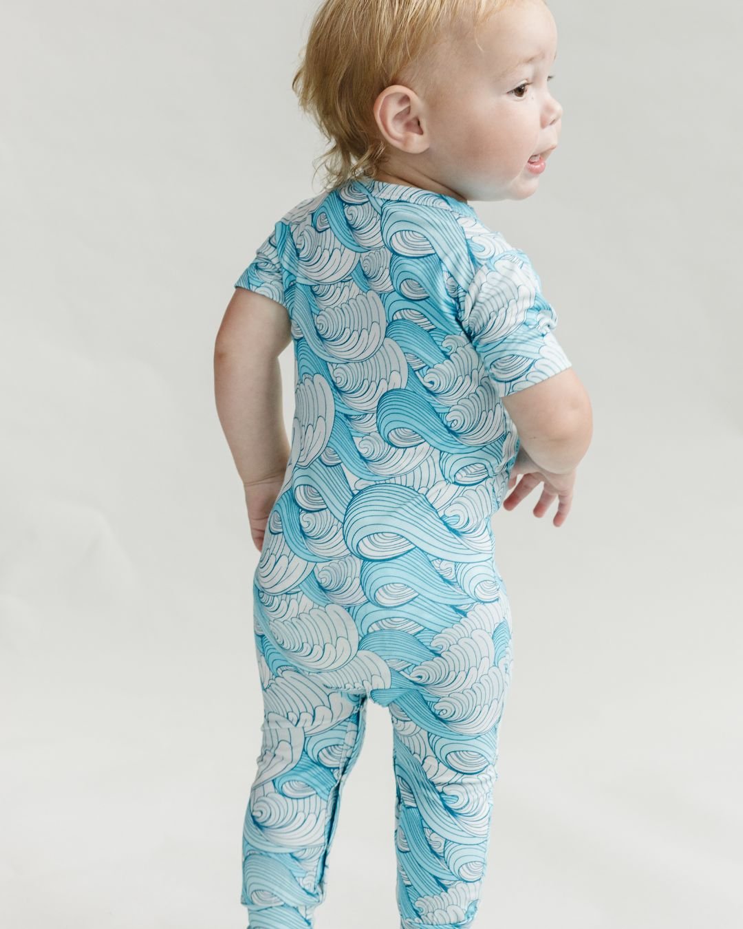 Lucky Panda Kids short sleeve romper in blue and white ocean waves design worn by a toddler boy who is standing with his back towards the camera. 