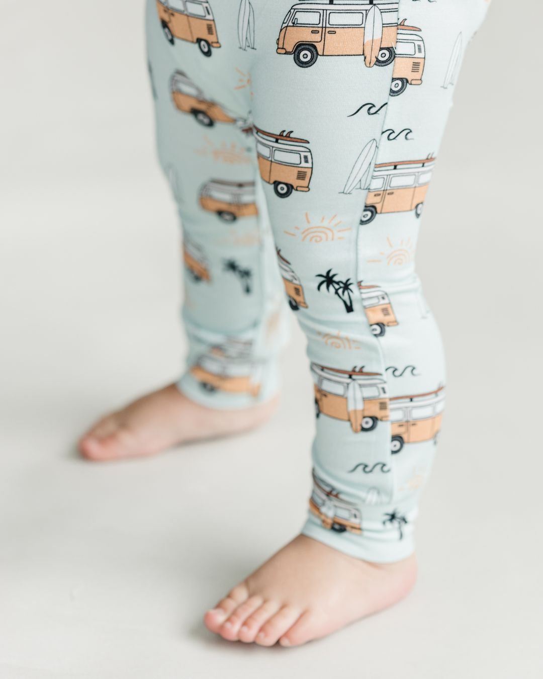 Close up leg shot of a baby boy standing up wearing a Lucky Panda Kids short sleeve cotton jumpsuit in light blue with orange surf van designs, palm trees, and surfboards.