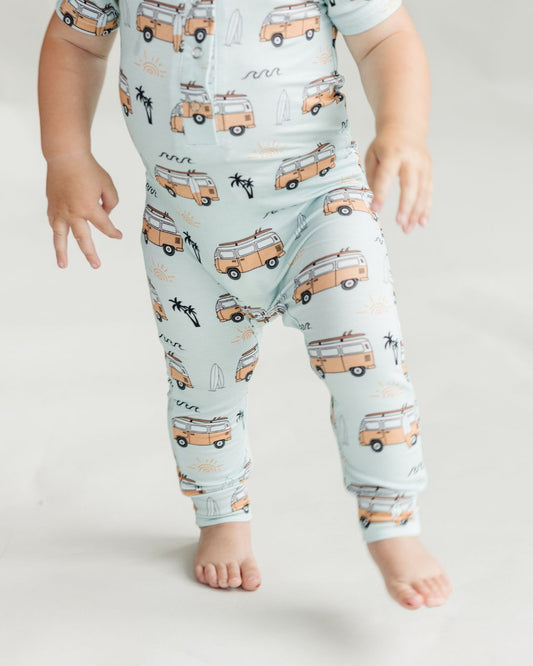 Baby boy standing up wearing a Lucky Panda Kids short sleeve cotton jumpsuit in light blue with orange surf van designs, palm trees, and surfboards.