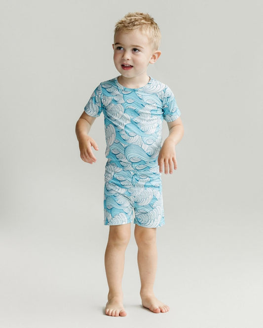 A little boy standing up wearing Lucky Panda Kids shorts set. It includes shorts and a t-shirt that are blue with white waves in the theme of california surf.