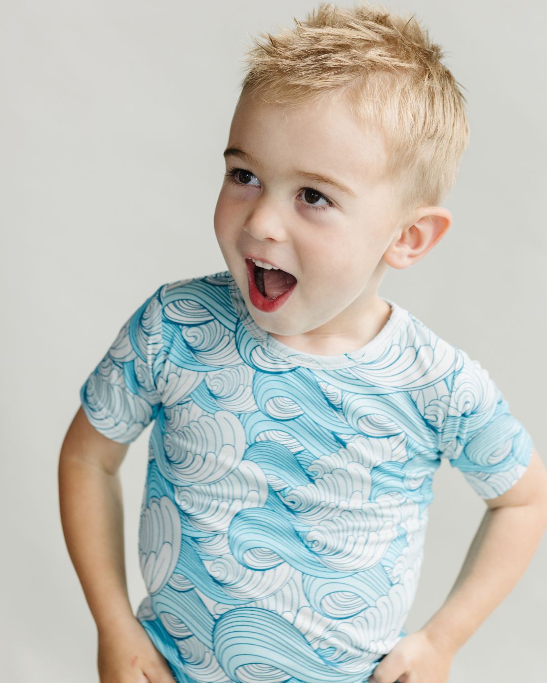 A little boy standing up wearing Lucky Panda Kids shorts set. It includes shorts and a t-shirt that are blue with white waves in the theme of california surf.