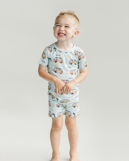 Little boy smiling and standing wearing the Lucky Panda Kids shorts set for babies and kids. He is wearing both a pair of shorts and a t-shirt that are blue with light orange surf vans and surf boards in the theme of california surf laid out on a blank background.