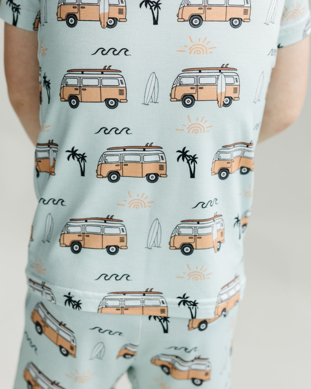 Close up shot from the back of a little boy standing wearing the Lucky Panda Kids shorts set for babies and kids. He is wearing both a pair of shorts and a t-shirt that are blue with light orange surf vans and surf boards in the theme of california surf laid out on a blank background.