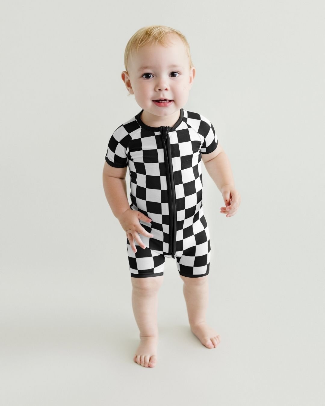 Toddler boy standing and wearing a Lucky Panda Kids bamboo and cotton black and white checkered short sleeve shortie romper.