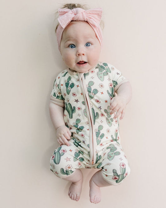 Infant baby girl laying down wearing a Lucky Panda Kids cream colored short sleeve shortie romper with green cactus designs, pink trim and pink flowers and stars laid out on a white background. 