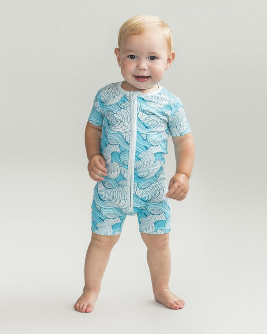 Toddler boy standing up wearing a Lucky Panda Kids short sleeve short romper made from bamboo with light blue, medium blue and white ocean wave designs. 