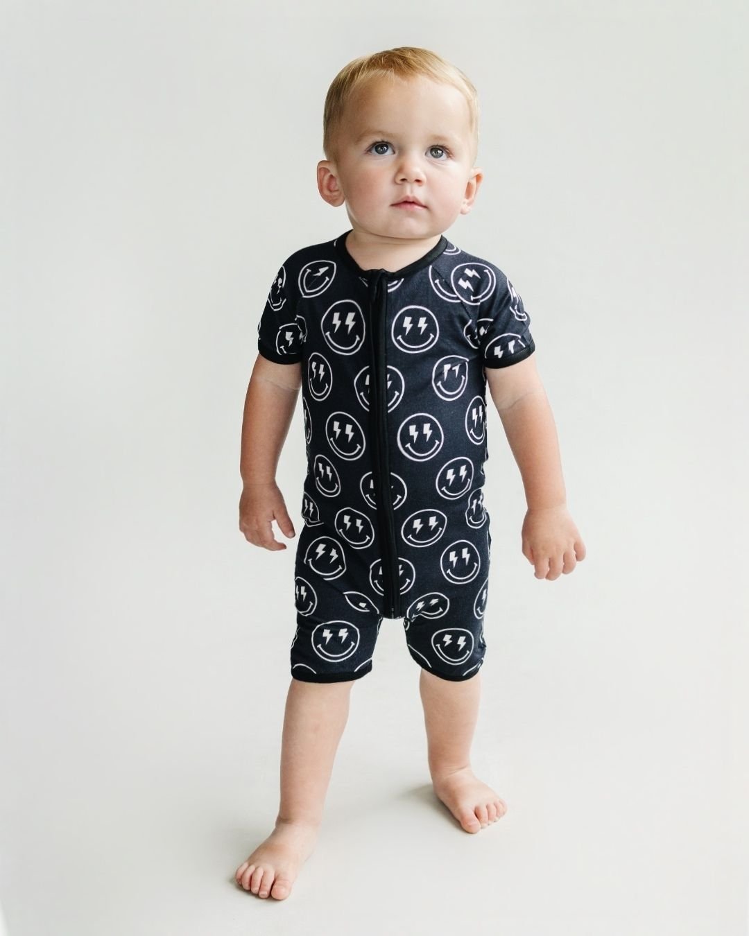 Toddler boy standing up wearing a Lucky Panda Kids black bamboo short sleeved shortie romper with white smiley face designs that have lightning bolts for eyes.