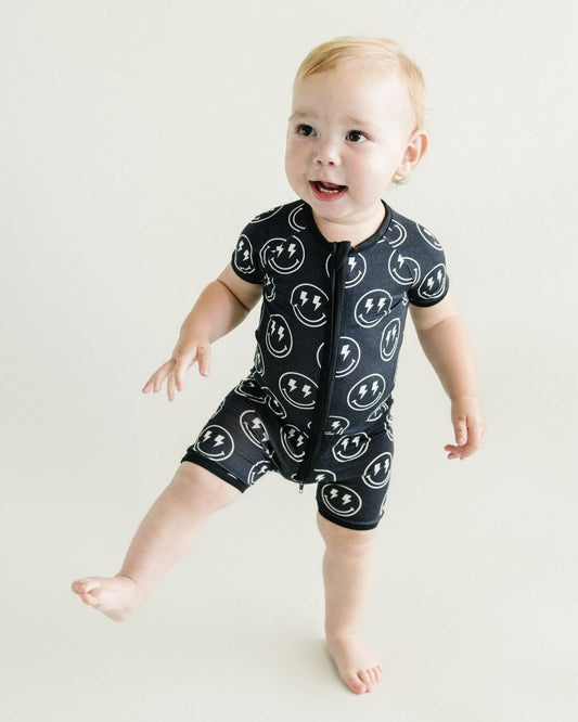 Toddler boy standing up wearing a Lucky Panda Kids black bamboo short sleeved shortie romper with white smiley face designs that have lightning bolts for eyes.