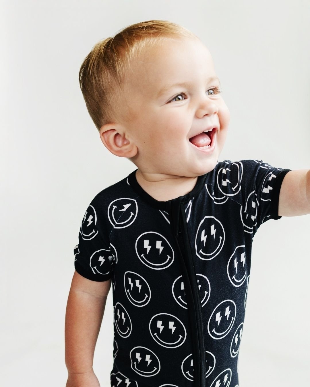 Toddler boy standing up wearing a Lucky Panda Kids black bamboo short sleeved shortie romper with white smiley face designs that have lightning bolts for eyes.