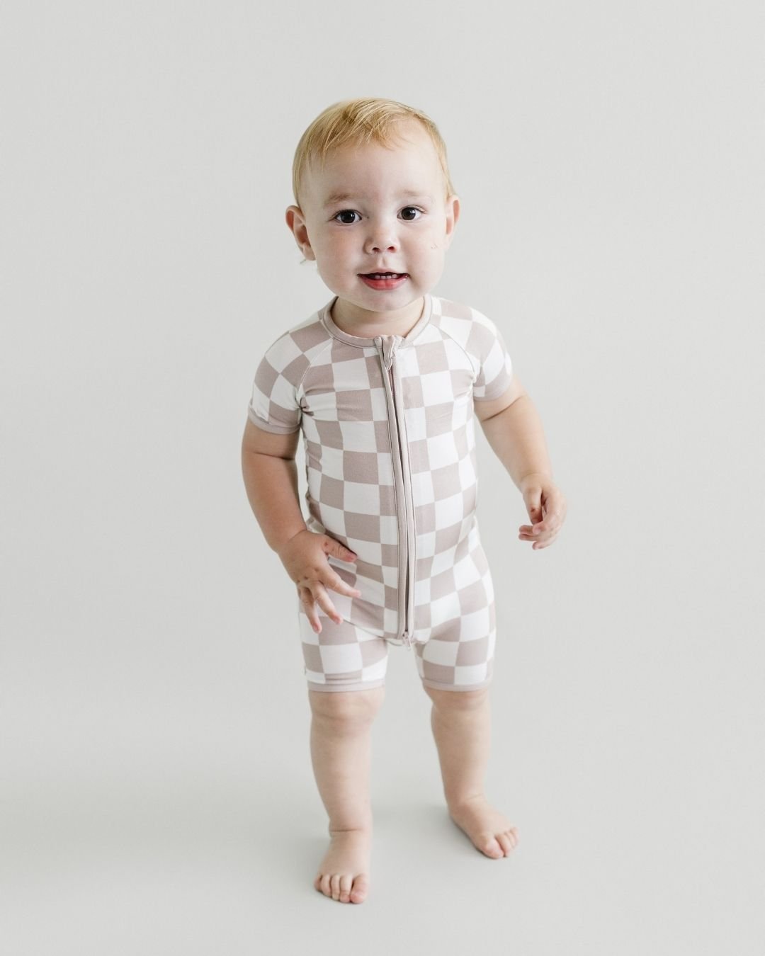 Toddler boy standing up wearing a Lucky Panda Kids short sleeve bamboo shortie romper in white and tan checkered design. 