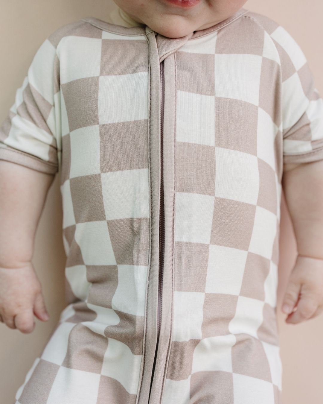 Close up shot of an infant baby laying down wearing a Lucky Panda Kids short sleeve bamboo shortie romper in white and tan checkered design. 