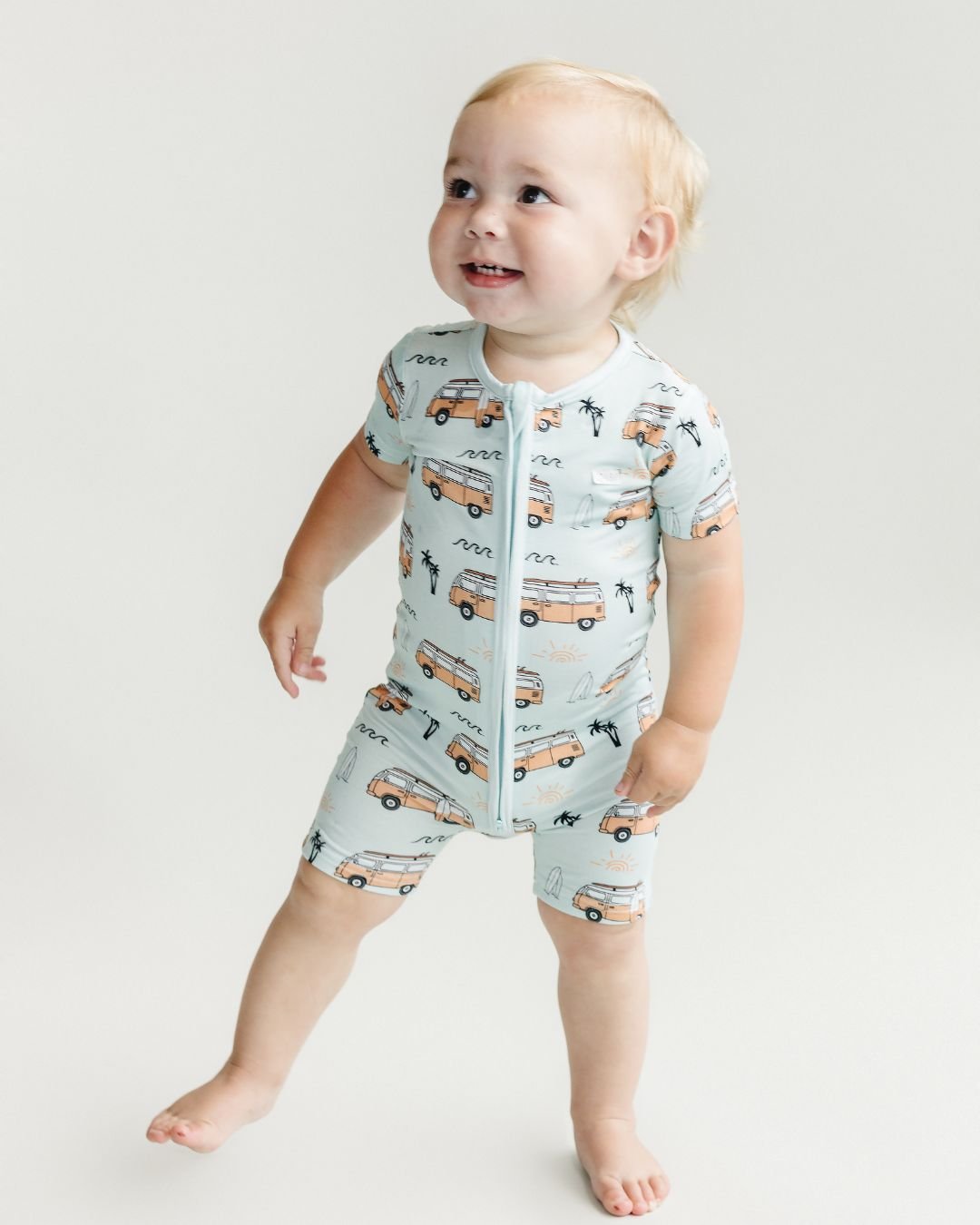 Toddler boy standing up wearing a Lucky Panda Kids blue bamboo short sleeved shortie romper with orange surf vans, white surfboards, palm trees and black wave designs. 