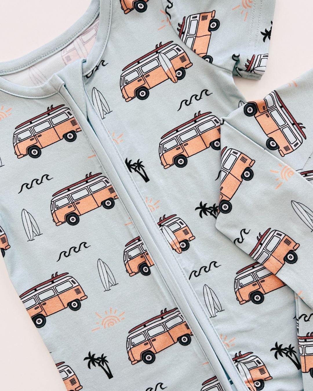 Close up design shot of a Lucky Panda Kids blue bamboo short sleeved shortie romper with orange surf vans, white surfboards, palm trees and black wave designs. 