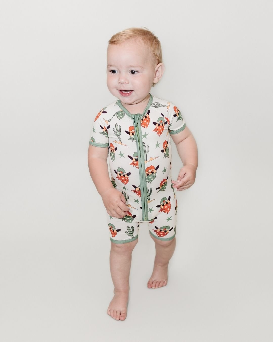 Toddler boy standing up wearing a Lucky Panda Kids short sleeve shortie romper. The design is white with sage green trimmings, cactus', stars and a cowboy with an orange starred bandana. 