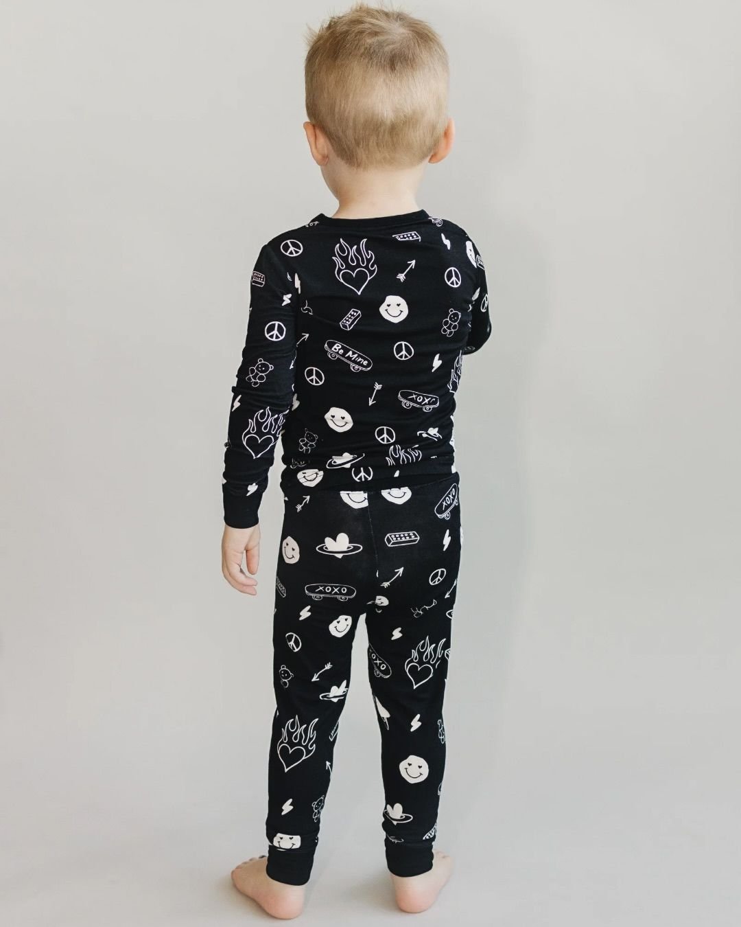  A boy standing up with his back facing the camera. He is wearing a Lucky Panda Kids two-piece long sleeve pajama set in black with white designs including smiley faces, hearts, arrows, and skateboards. 