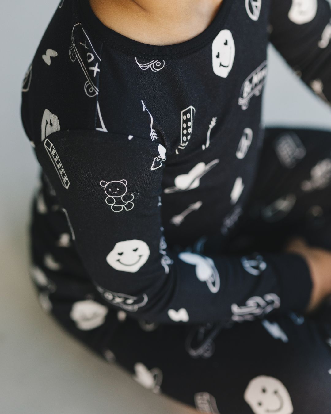 Close up shot of a kid wearing a Lucky Panda Kids two-piece long sleeve pajama set in black with white designs including smiley faces, hearts, arrows, and skateboards. 