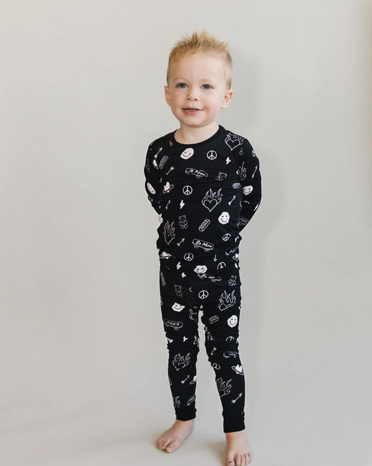  A boy standing up and wearing a Lucky Panda Kids two-piece long sleeve pajama set in black with white designs including smiley faces, hearts, arrows, and skateboards. 