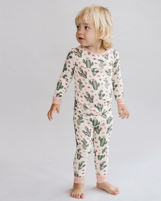 A young boy standing up wearing a two piece pajama set in cream color with light pink trim, green cactus designs with small pink flowers and stars. The pj's are part of the viscose bamboo collection from Lucky Panda Kids. 