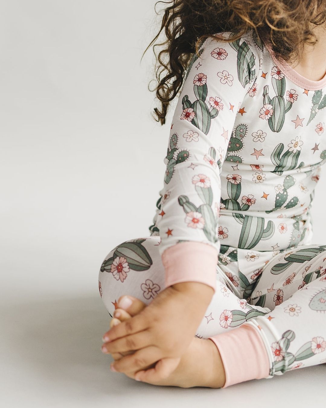 Close up body shot of a young girl sitting, wearing a two piece pajama set in cream color with light pink trim, green cactus designs with small pink flowers and stars. The pj's are part of the viscose bamboo collection from Lucky Panda Kids. 