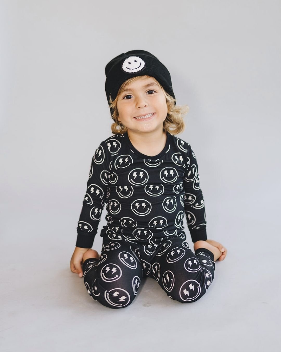 A young boy kneeling wearing a two piece black viscose bamboo pajama set from Lucky Panda Kids with white smiley faces and lightning bolts for eyes on the pj's. 