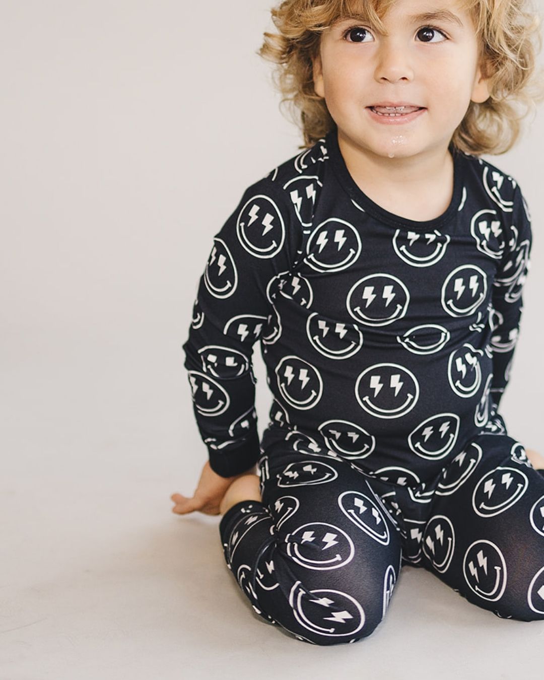 A young boy kneeling, wearing a two piece black viscose bamboo pajama set from Lucky Panda Kids with white smiley faces and lightning bolts for eyes on the pj's. 