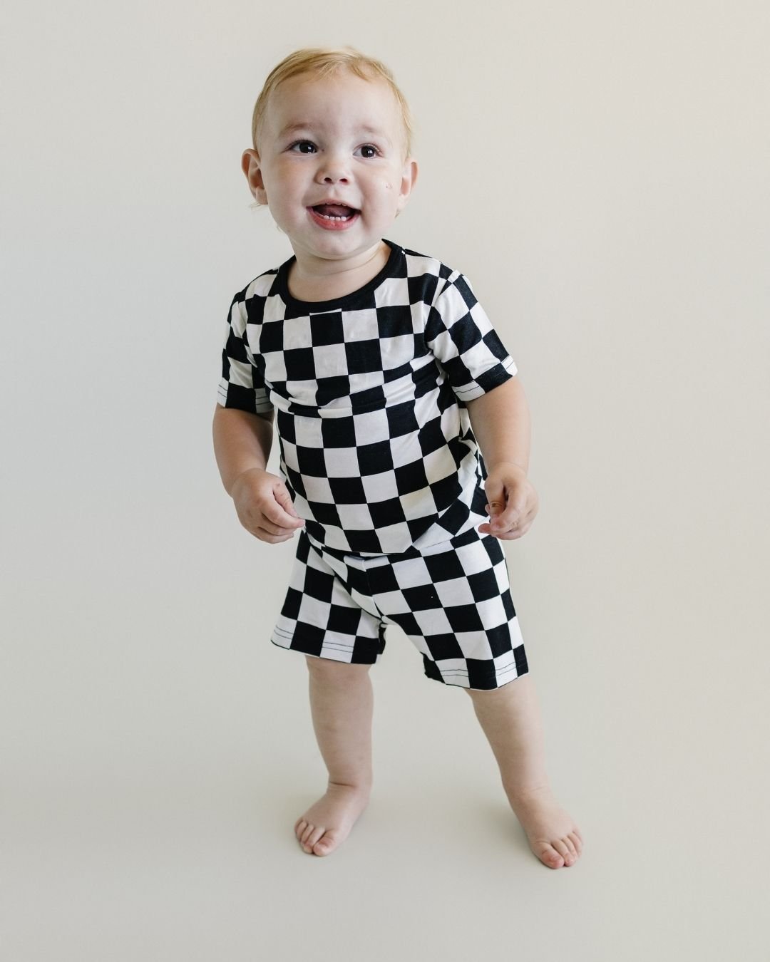 A toddler boy standing up smiling and wearing a Lucky Panda Kids two-piece soft viscose bamboo pajama shorts set in a white and black checkered print.