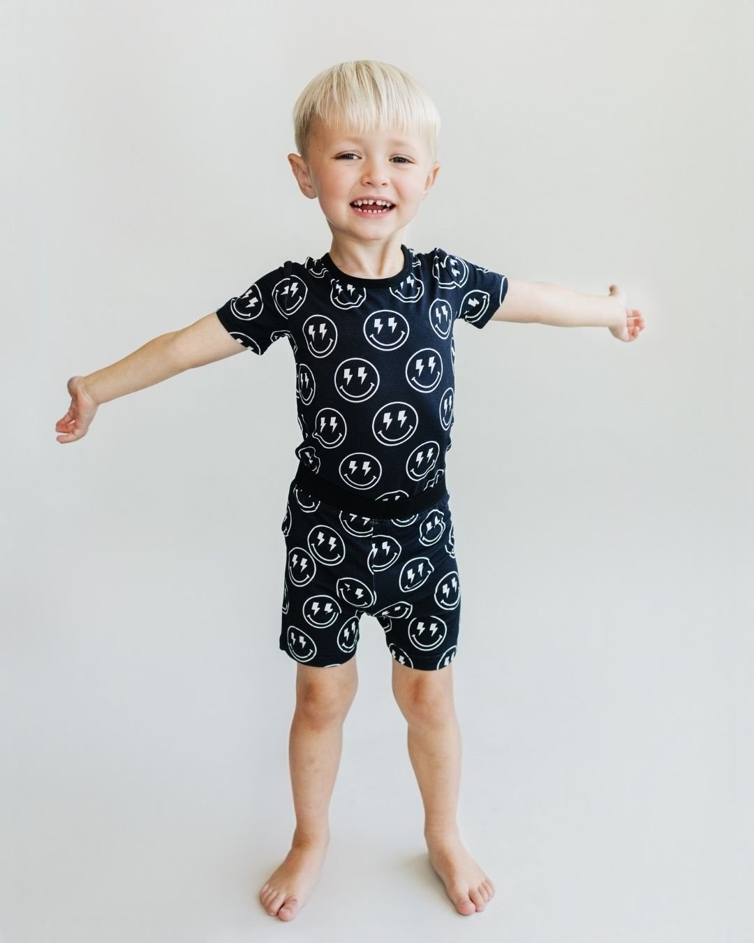 A young boy standing up with his arms spread wide. He is wearing the Lucky Panda Kids two-piece pajama set. It is black with white smiley faces that have lightning bolts for eyes. 