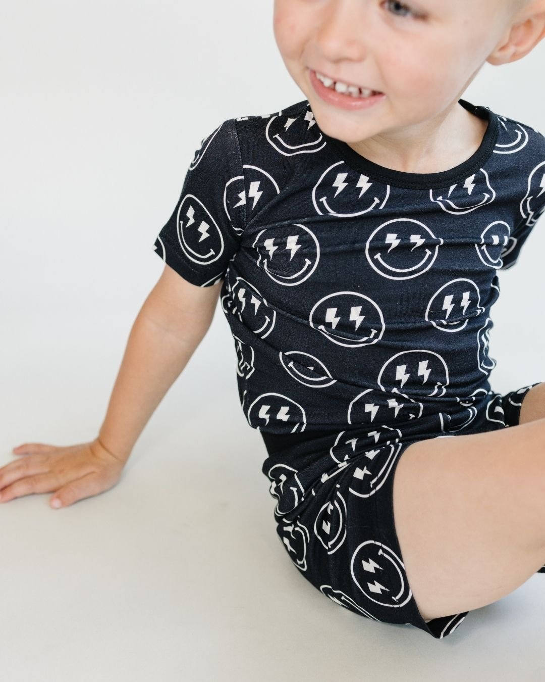 A young boy sitting on the floor. He is wearing the Lucky Panda Kids two-piece pajama set. It is black with white smiley faces that have lightning bolts for eyes. 