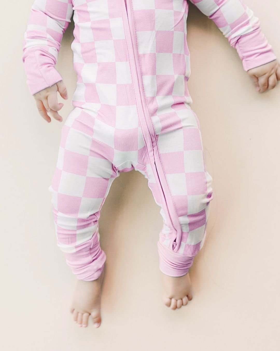 Close up detailed headless shot of an infant girl laying down wearing a Lucky Panda Kids soft viscose bamboo zip up pajama in a white and light pink checkered pattern.