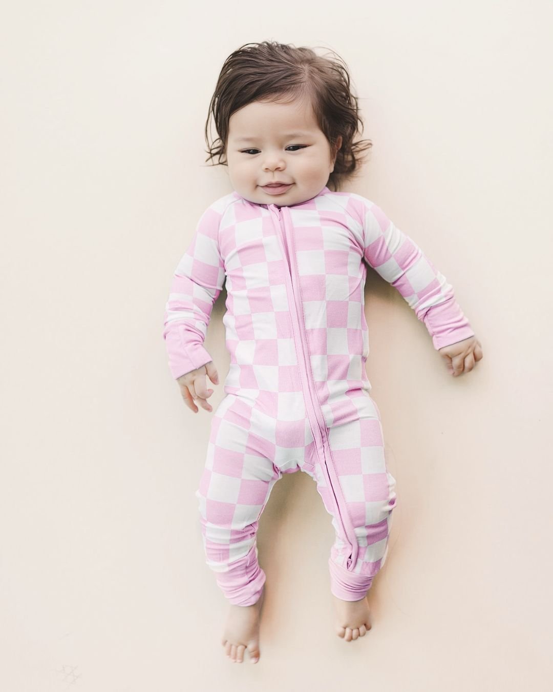 An infant girl laying down wearing a Lucky Panda Kids soft viscose bamboo zip up pajama in a white and light pink checkered pattern.