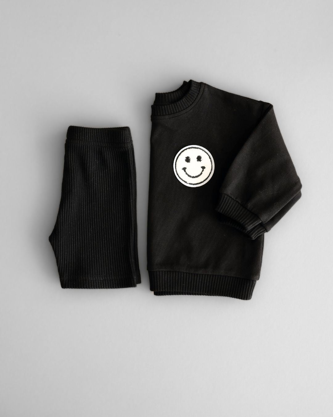 A white background featuring a folded black cotton Lucky Panda Kids "Biker Set" which includes ribbed cotton shorts and a sweatshirt with a fuzzy white smiley face emblem on the top right.