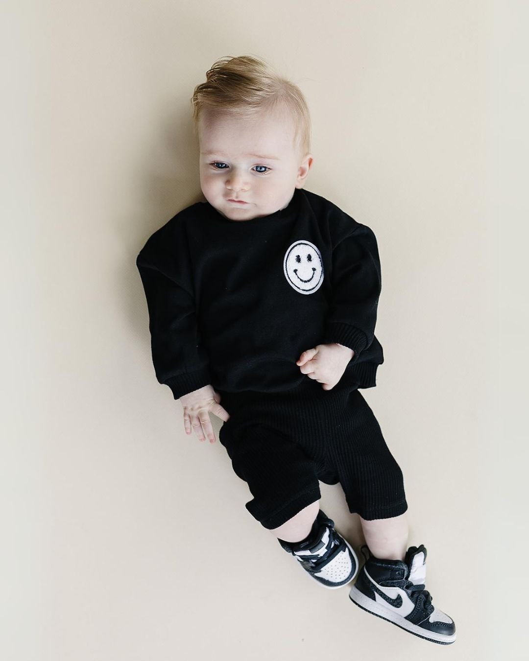 A baby boy laying down wearing a black cotton Lucky Panda Kids "Biker Set" which includes ribbed cotton shorts and a sweatshirt with a fuzzy white smiley face emblem on the top right.