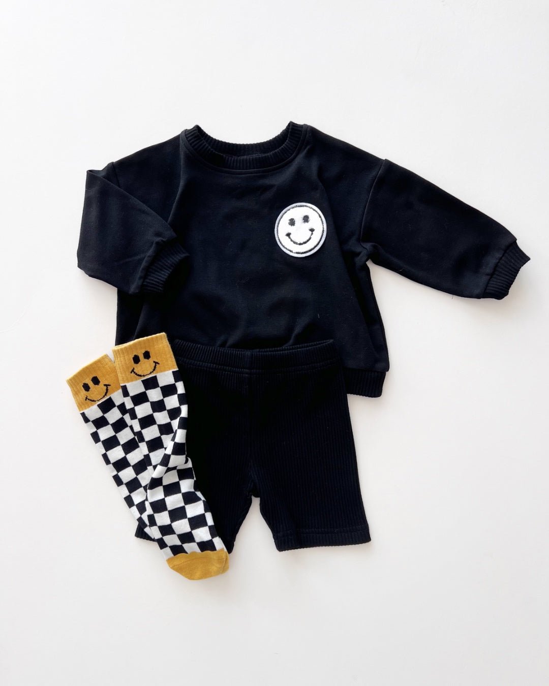 A white background featuring a black cotton Lucky Panda Kids "Biker Set" which includes ribbed cotton shorts and a sweatshirt with a fuzzy white smiley face emblem on the top right.