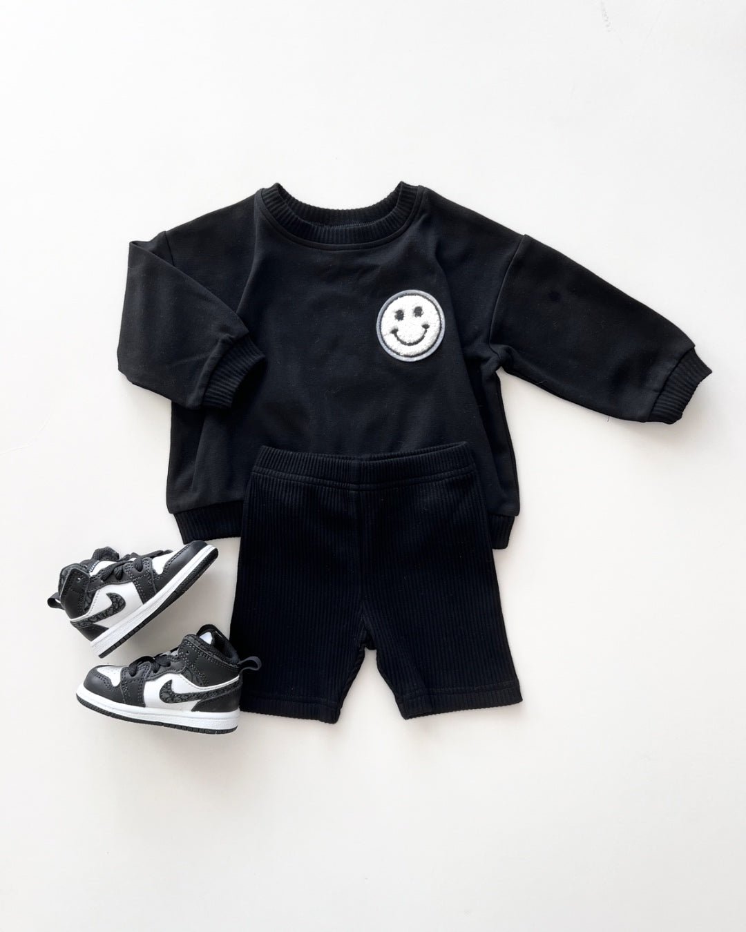 A white background featuring a black cotton Lucky Panda Kids "Biker Set" which includes ribbed cotton shorts and a sweatshirt with a fuzzy white smiley face emblem on the top right.