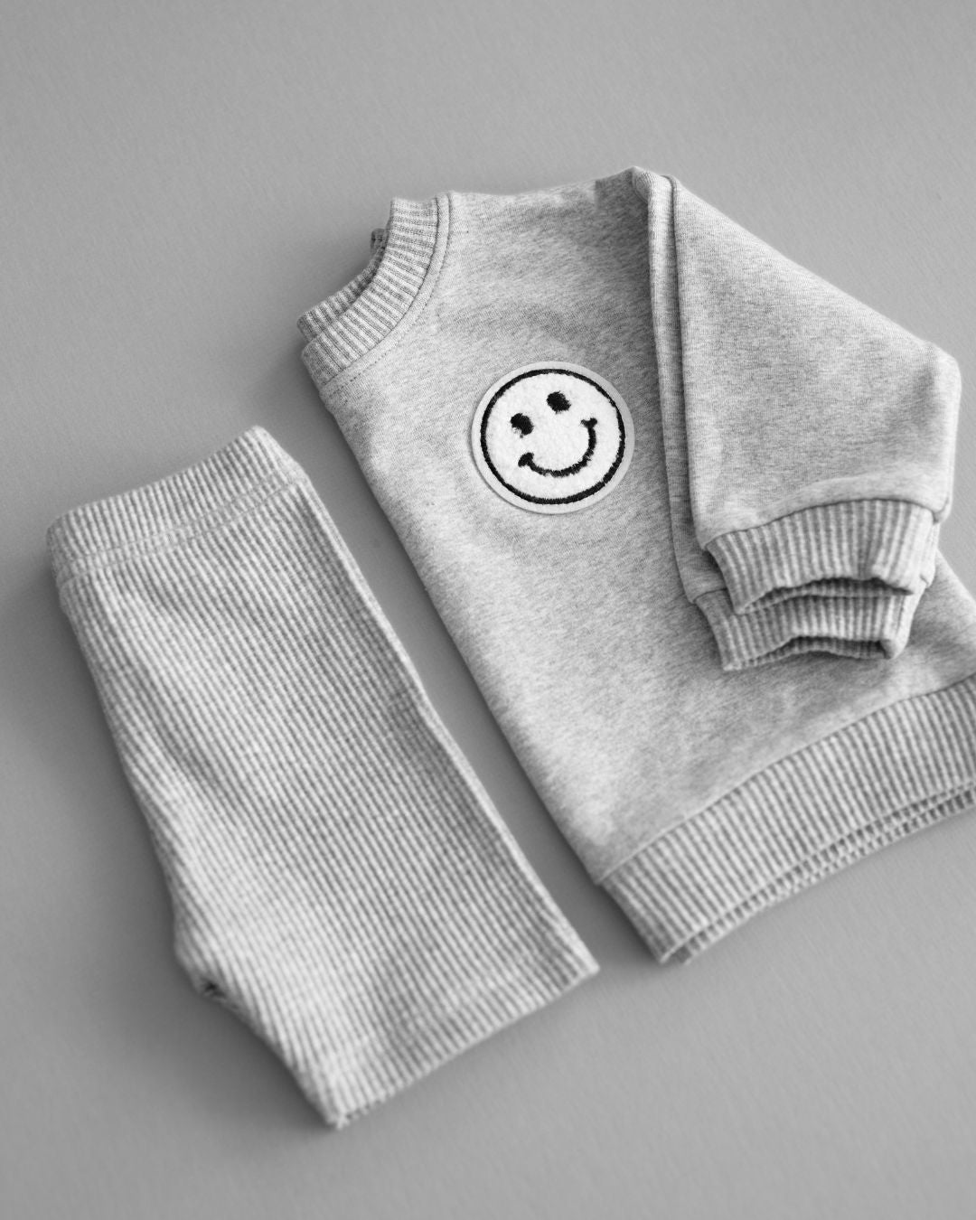 A gray background with a folded light gray cotton Lucky Panda Kids "Biker Set" which includes ribbed cotton shorts and a sweatshirt with a fuzzy white smiley face emblem on the top right.