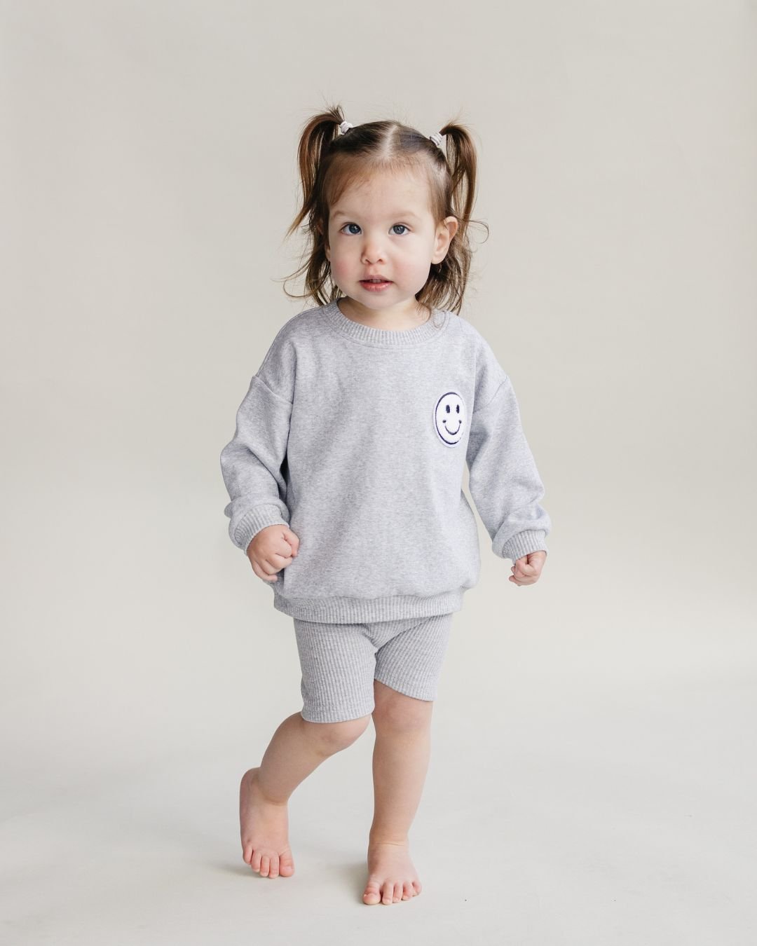 A toddler girl standing up. Se is wearing a light gray cotton Lucky Panda Kids "Biker Set" which includes ribbed cotton shorts and a sweatshirt with a fuzzy white smiley face emblem on the top right.