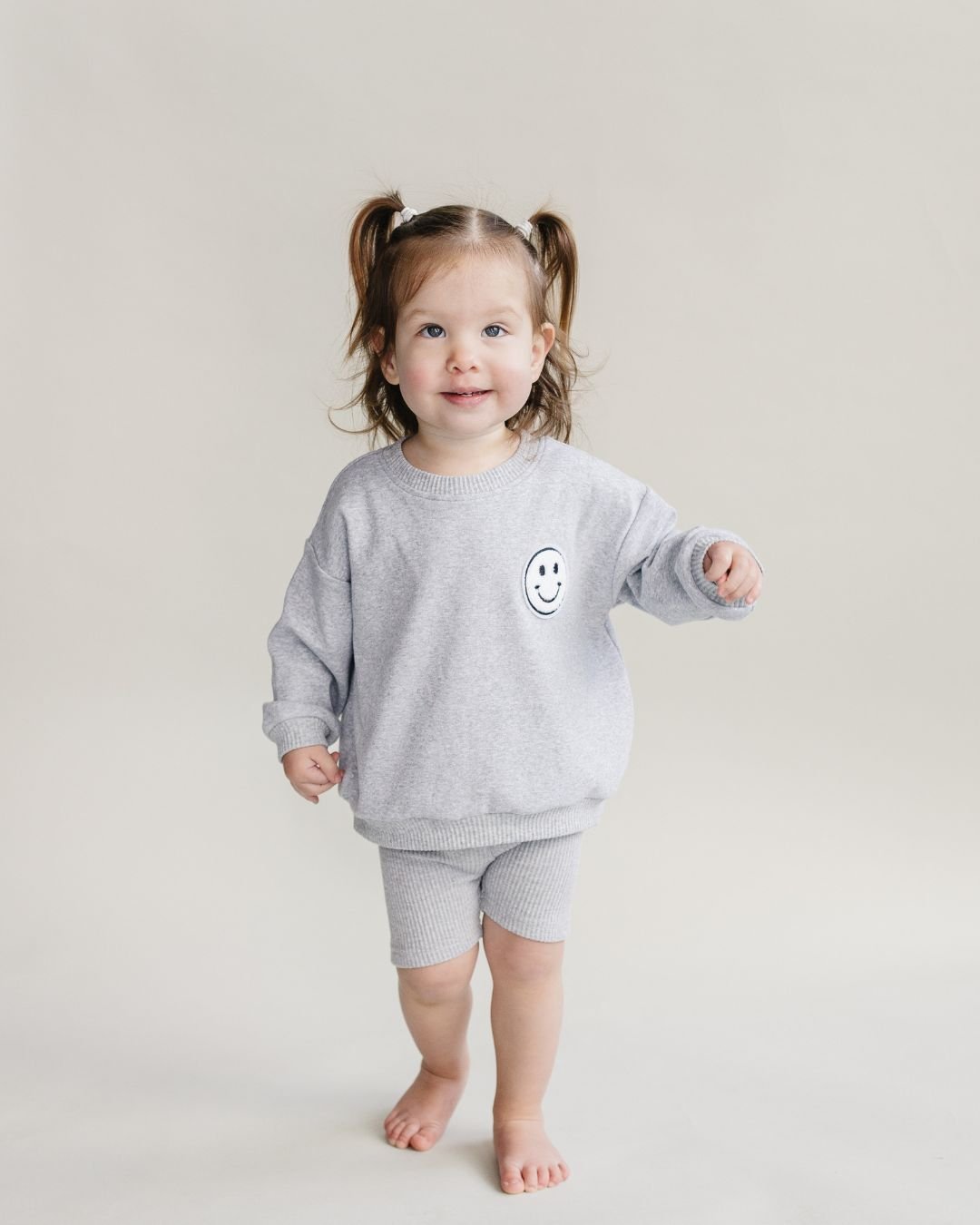 A toddler girl standing up smiling. She is wearing a light gray cotton Lucky Panda Kids "Biker Set" which includes ribbed cotton shorts and a sweatshirt with a fuzzy white smiley face emblem on the top right.