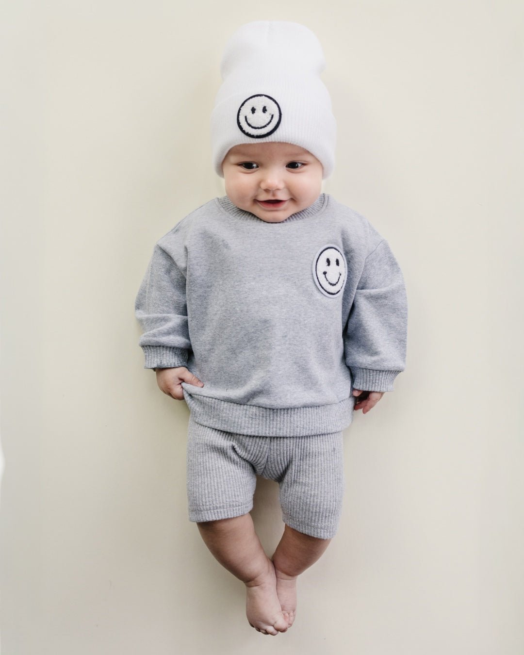 An infant boy laying down and smiling. He is wearing a light gray cotton Lucky Panda Kids "Biker Set" which includes ribbed cotton shorts and a sweatshirt with a fuzzy white smiley face emblem on the top right.