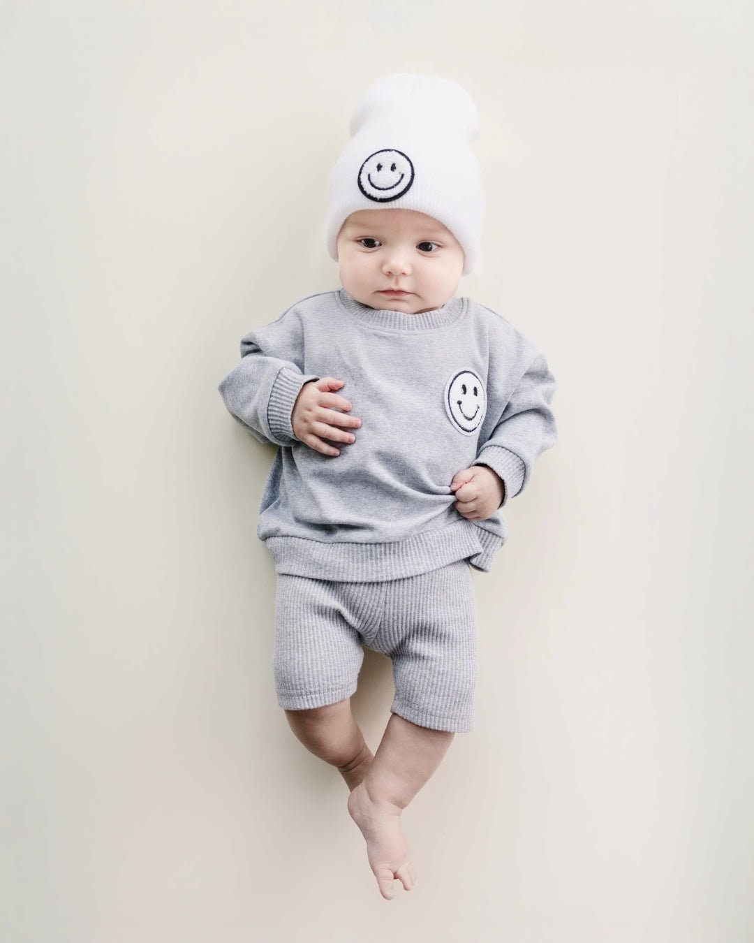 An infant boy laying down. He is wearing a light gray cotton Lucky Panda Kids "Biker Set" which includes ribbed cotton shorts and a sweatshirt with a fuzzy white smiley face emblem on the top right.