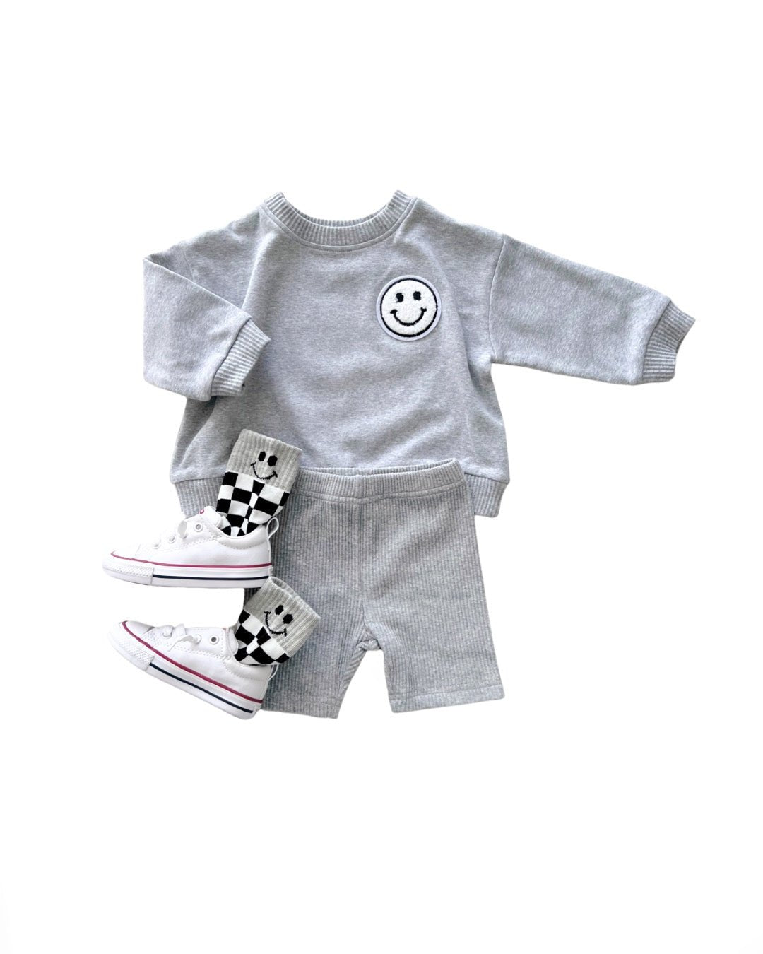 A white background with a laid out light gray cotton Lucky Panda Kids "Biker Set" which includes ribbed cotton shorts and a sweatshirt with a fuzzy white smiley face emblem on the top right. It is paired with checkered socks and white baby converse as accessories. 