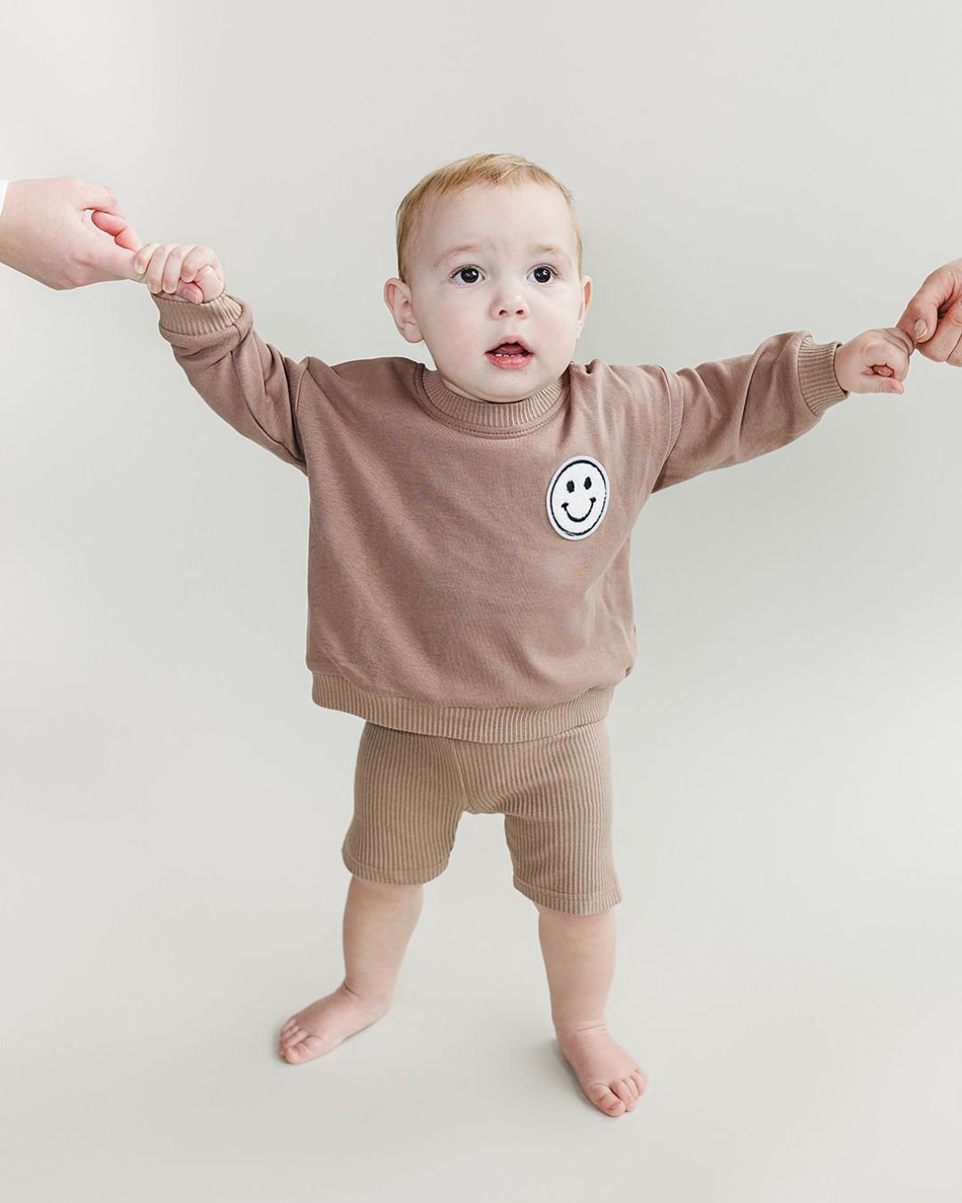 Baby boy standing up wearing a light brown cotton biker shorts set from Lucky Panda Kids. The shorts are ribbed cotton, and the sweatshirt has a white smiley face emblem on the top right.