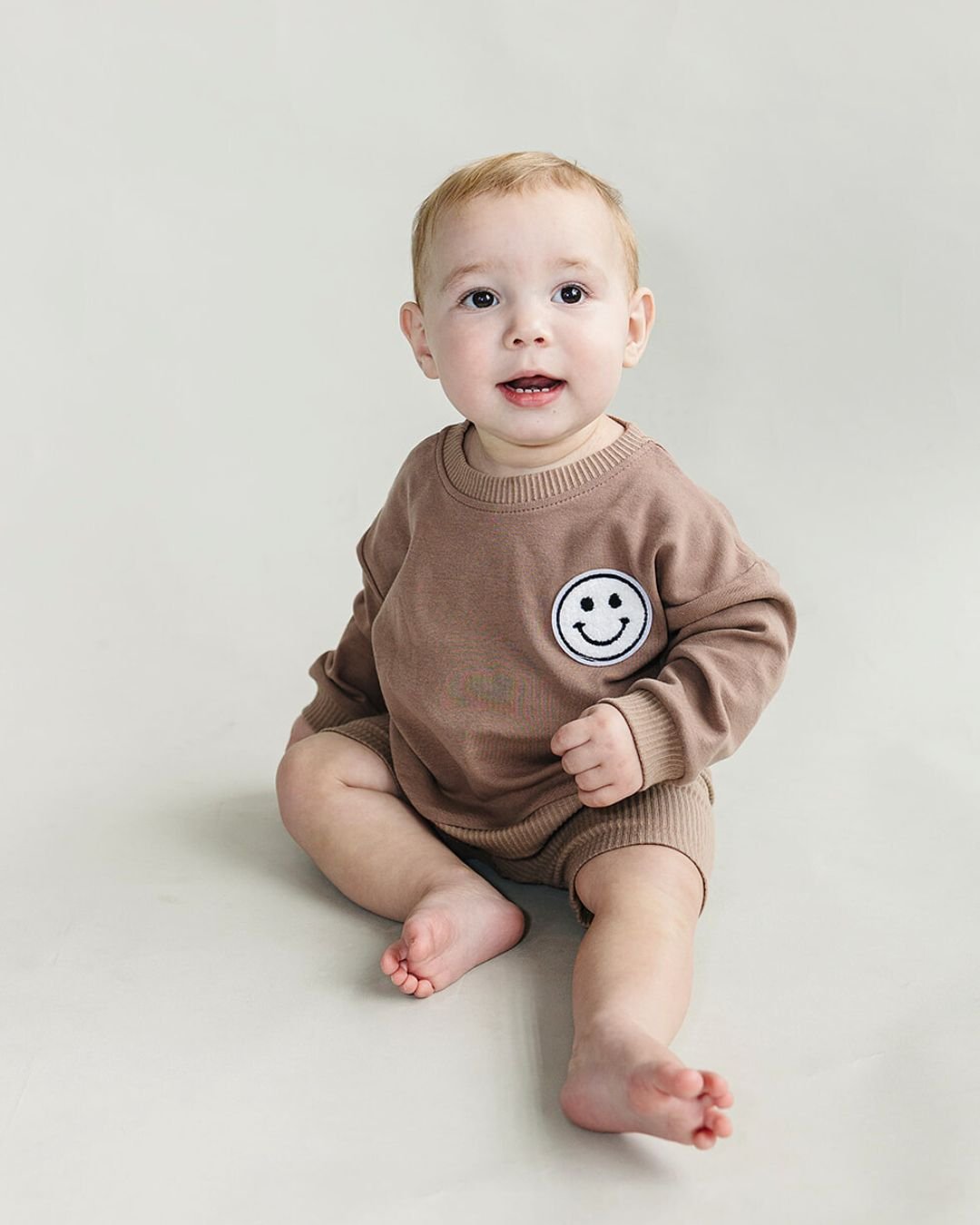 Baby boy sitting up wearing a light brown cotton biker shorts set from Lucky Panda Kids. The shorts are ribbed cotton, and the sweatshirt has a white smiley face emblem on the top right.