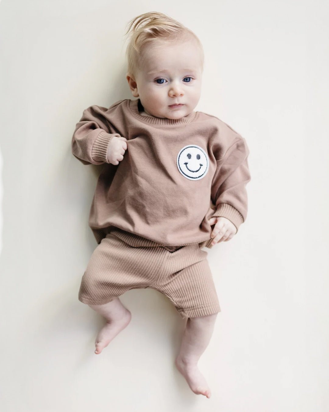 Baby boy laying on his back wearing a light brown cotton biker shorts set from Lucky Panda Kids. The shorts are ribbed cotton, and the sweatshirt has a white smiley face emblem on the top right.