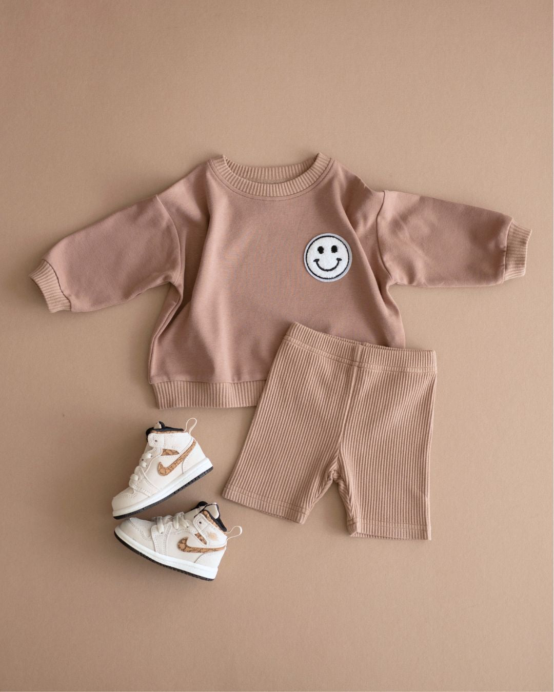 Brown background featuring a light brown cotton biker shorts set from Lucky Panda Kids. The shorts are ribbed cotton, and the sweatshirt has a white smiley face emblem on the top right.