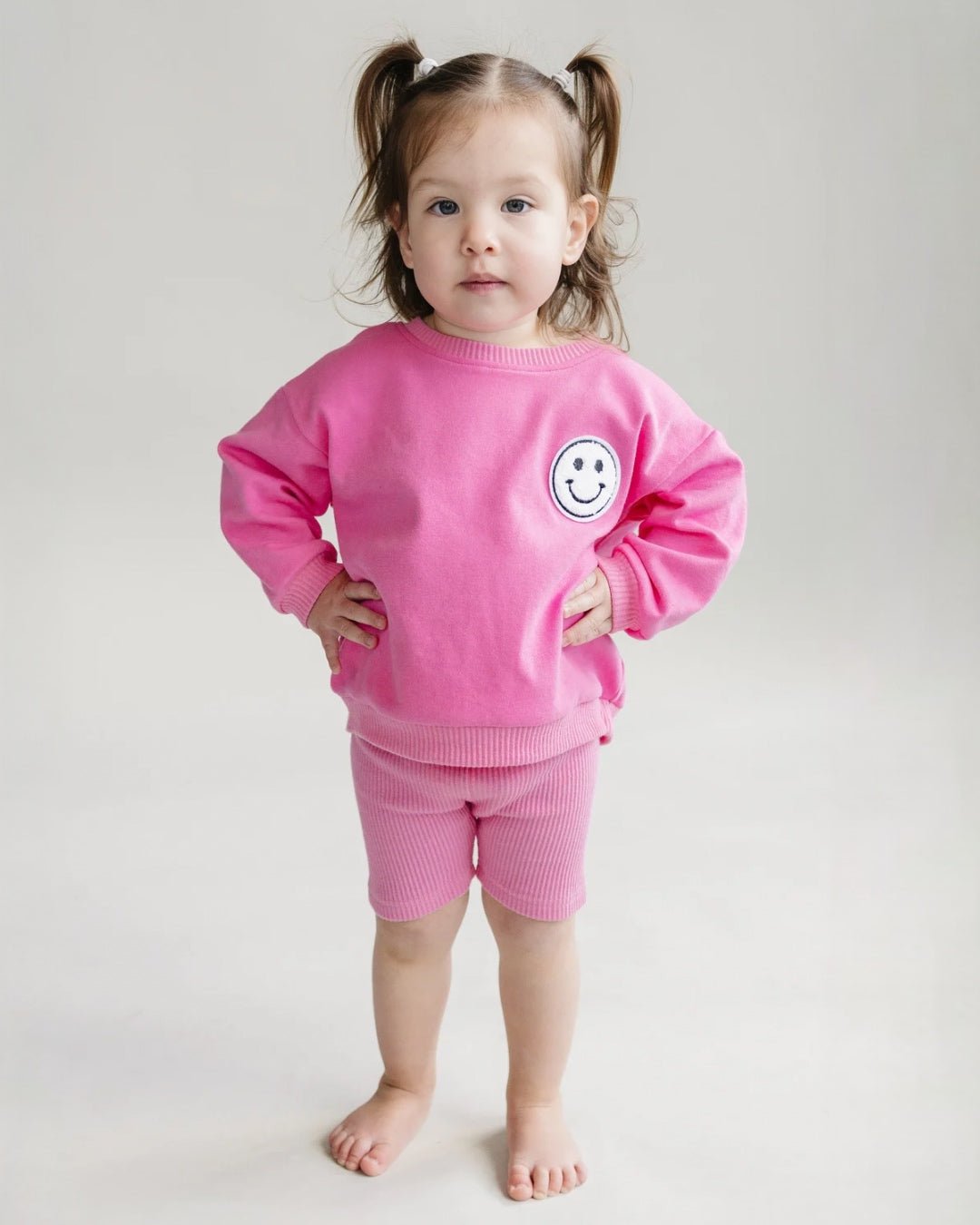 A toddler girl standing up wearing a bright pink cotton Lucky Panda Kids "Biker Set" which includes ribbed cotton shorts and a sweatshirt with a fuzzy white smiley face emblem on the top right.